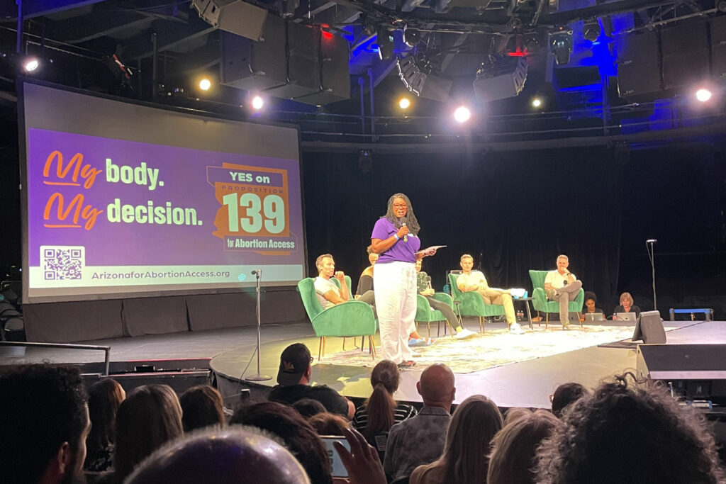 Chris Love on stage with Pod Save America in front of a large yes on prop 139 My Body, My Decision sign in front of a crowd in Phoenix, AZ